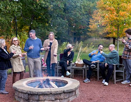 A group of friends gathers outdoors around a stone fire pit, toasting with drinks on a fall evening.