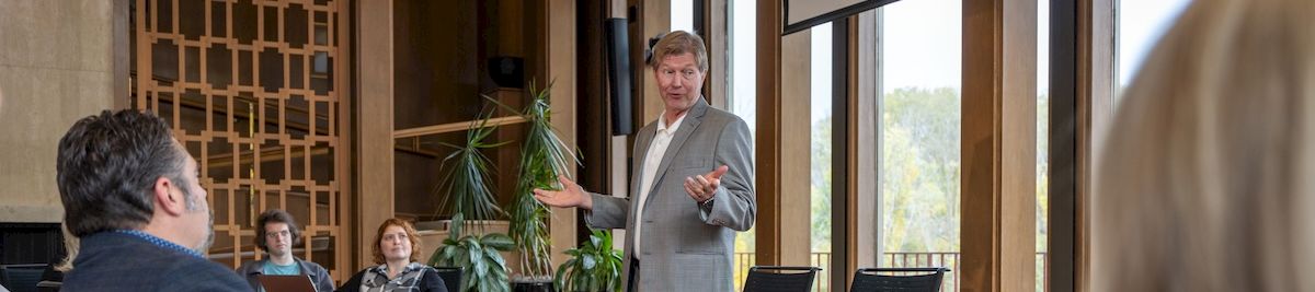 A business meeting in a conference room, with a presenter gesturing to colleagues seated around a table.