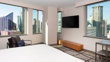A modern hotel room with a bed, armchair, wall-mounted TV, and large windows showcasing a cityscape view ending the sentence.
