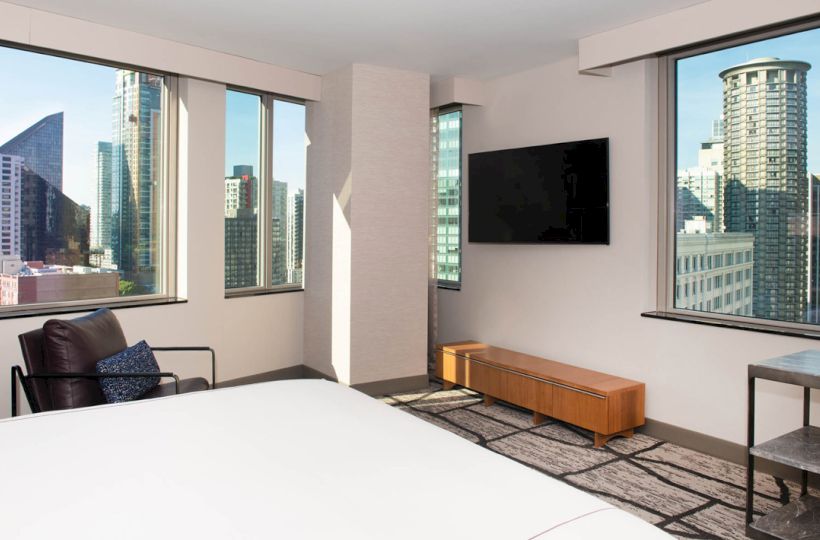 A modern hotel room with a bed, armchair, wall-mounted TV, and large windows showcasing a cityscape view ending the sentence.