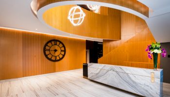 The image shows a modern lobby with wooden accents, a large clock, marble reception desk, and decorative ceiling lights.