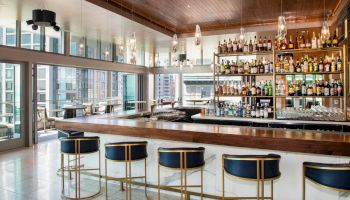 A modern bar with stylish seating, a variety of bottles displayed, and large windows offering a city view completes the scene.