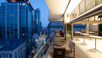 A rooftop terrace with tables and chairs overlooks a cityscape with tall buildings at dusk, featuring modern architecture and lighting.