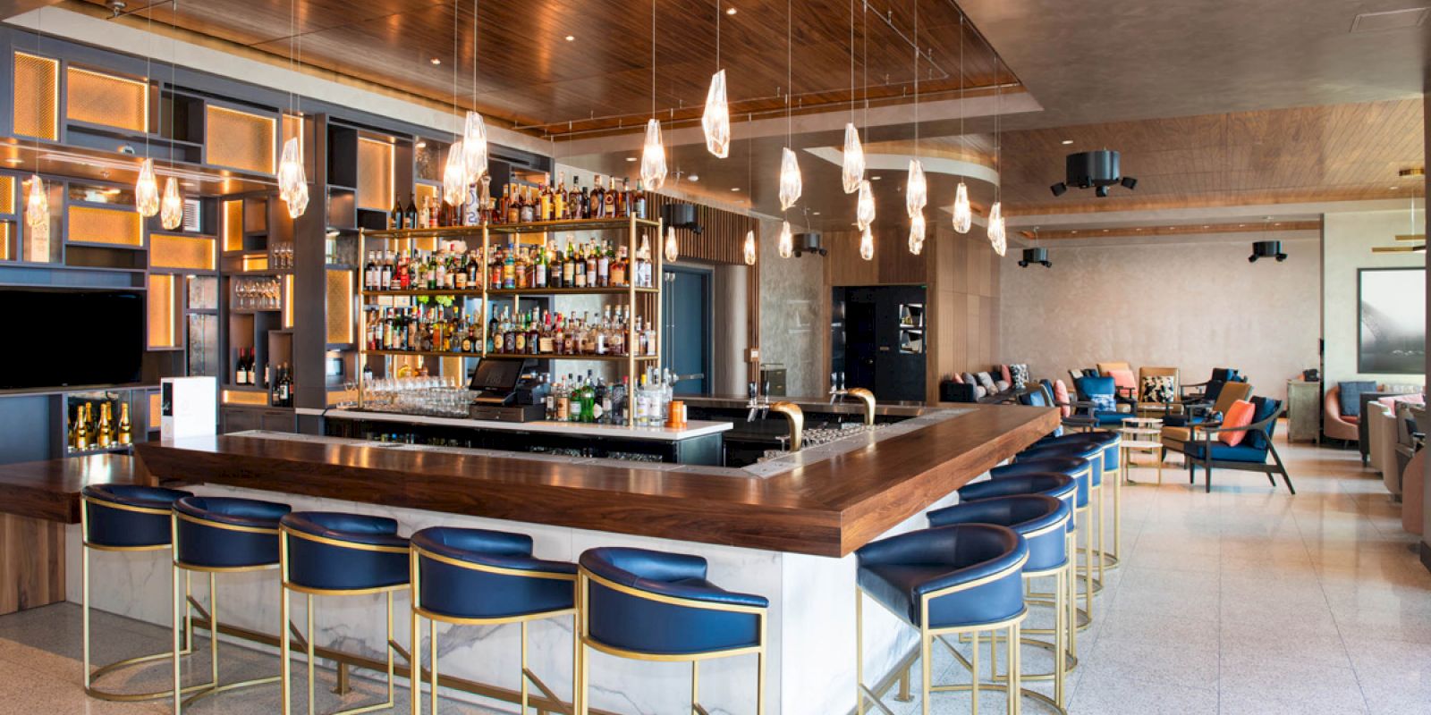 The image shows a modern bar with a central counter, stylish bar stools, a well-stocked display, and cozy seating in a sophisticated setting.