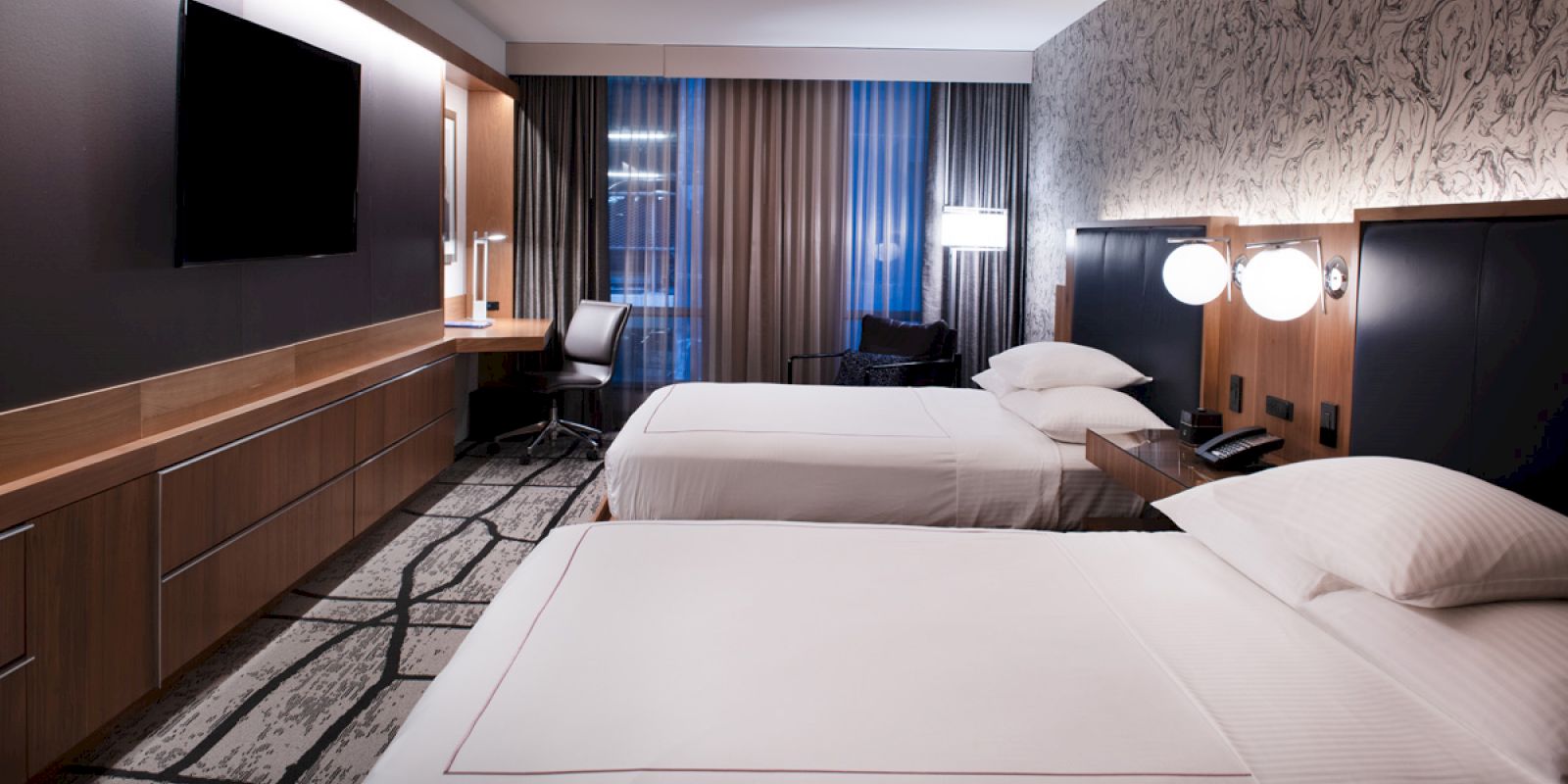 The image shows a modern hotel room with two beds, a wall-mounted TV, a desk, and a window with curtains.