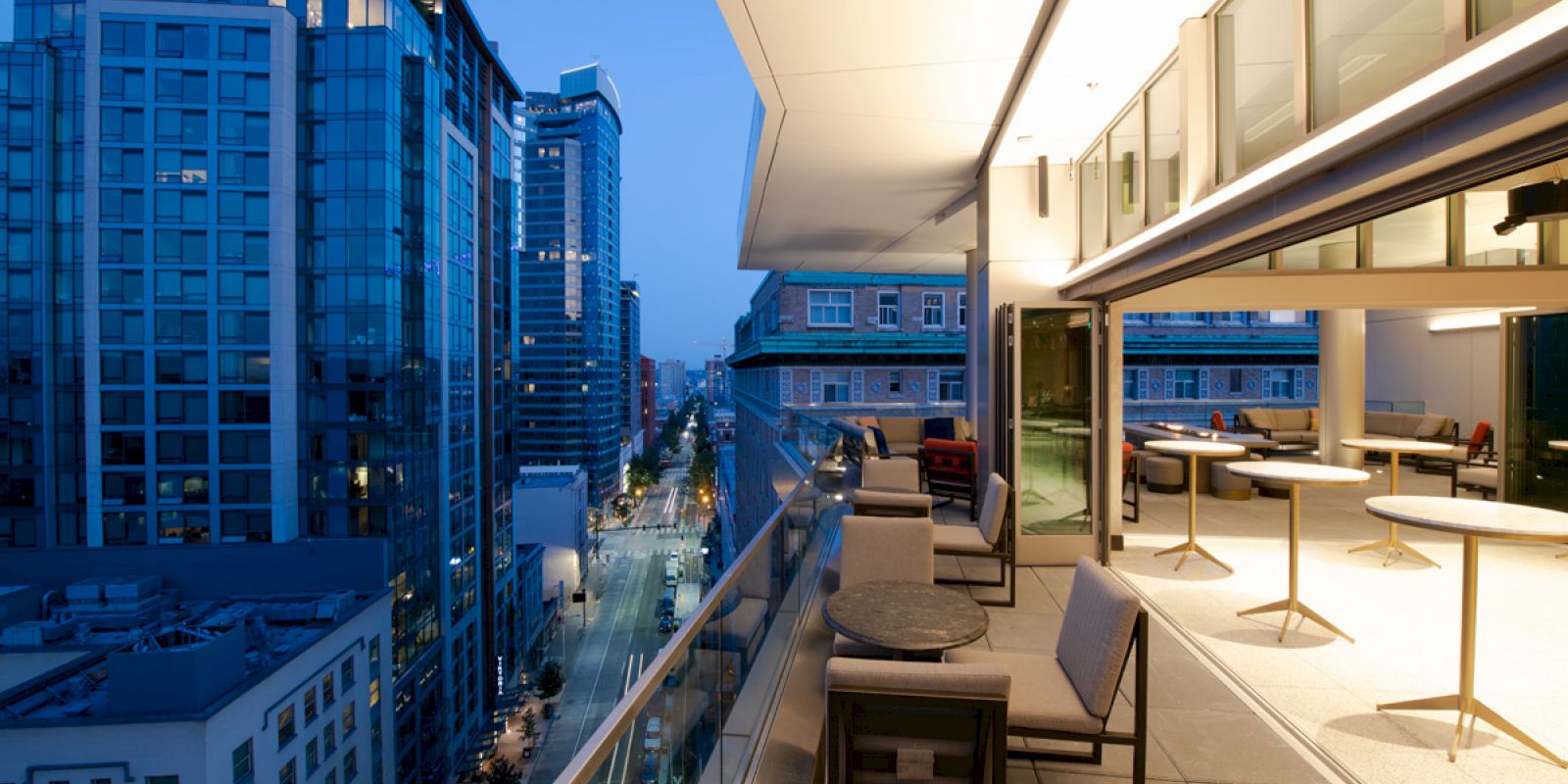 A modern cityscape at dusk with a balcony view featuring outdoor seating, overlooking a street lined with tall buildings.
