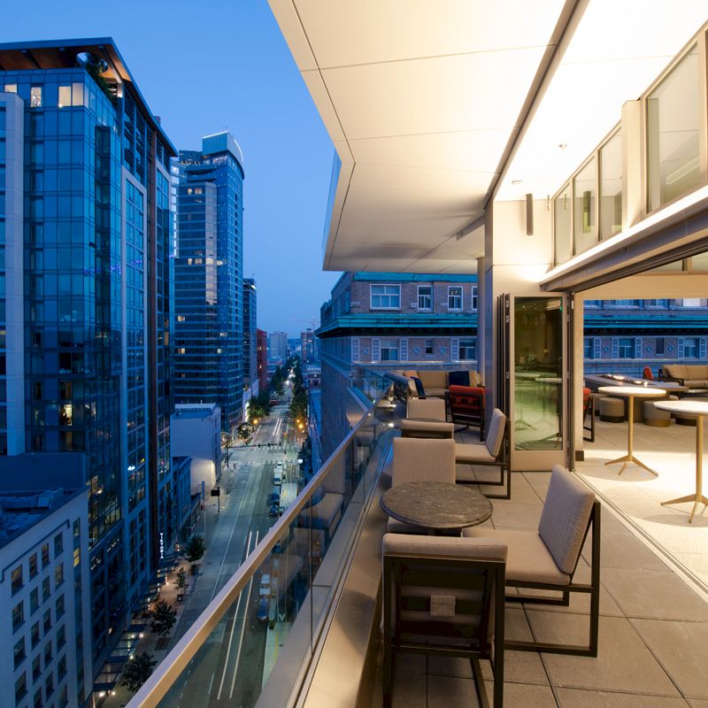A modern rooftop terrace with seating overlooks a cityscape of tall buildings at dusk, featuring open indoor and outdoor spaces.