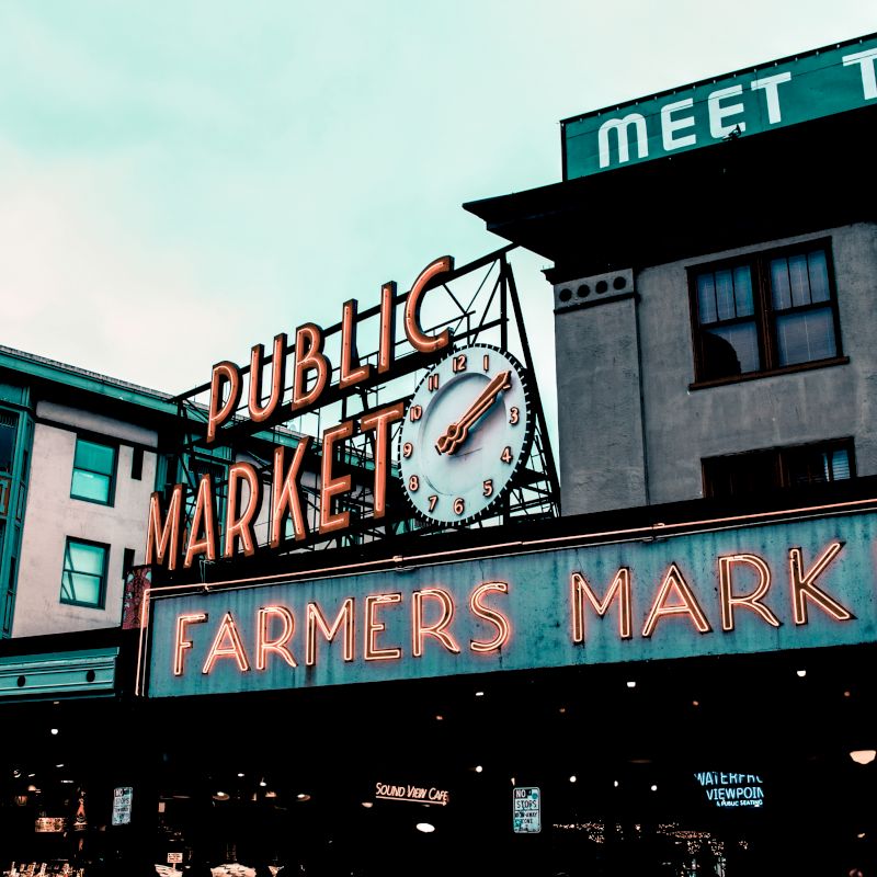 The image shows the iconic entrance sign for a public and farmers market, with a clock and surrounding buildings in the background.