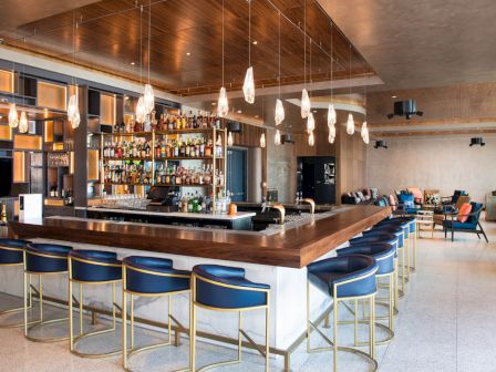 The image shows a modern bar with a central counter, stylish chairs, hanging lights, and shelves stocked with various bottles.
