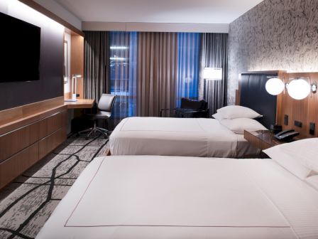 A modern hotel room with two beds, a TV, a work desk with chair, lamps, and decorative wallpaper, creating a cozy atmosphere.