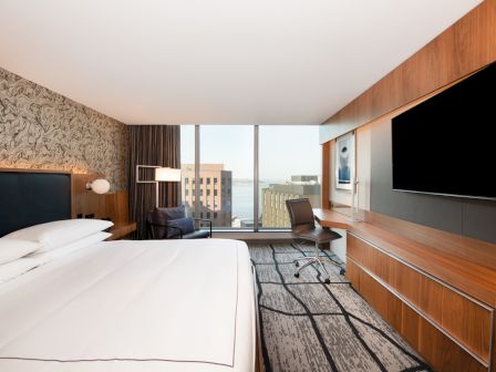 A modern hotel room with a bed, desk, chair, large TV, and view of buildings through a window.