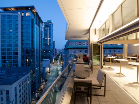 A modern cityscape at dusk with a balcony view, featuring tall buildings, glass railings, tables, and chairs overlooking the street below.