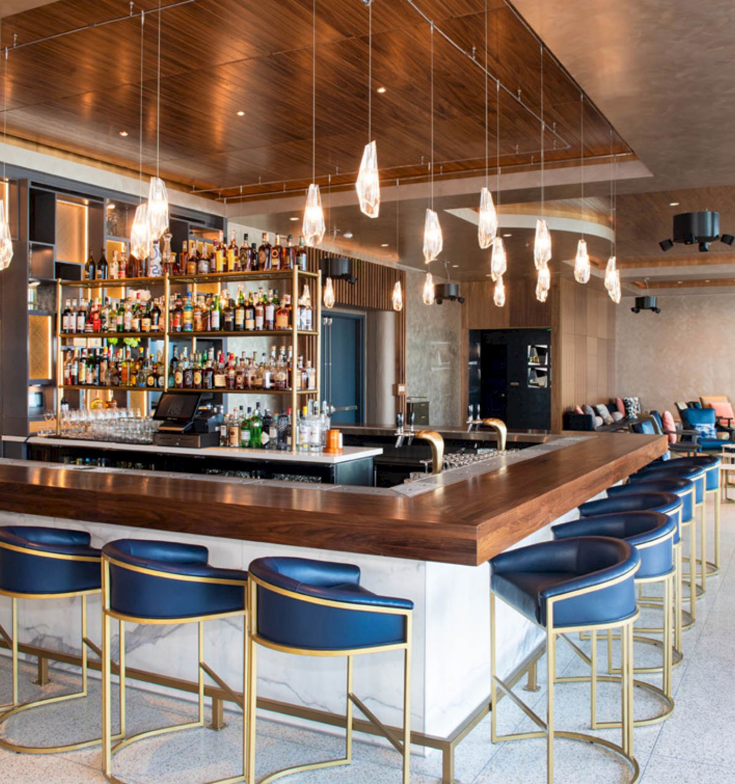 A modern bar with blue chairs, overhead lights, and a well-stocked liquor shelf in an open, stylish lounge setting.