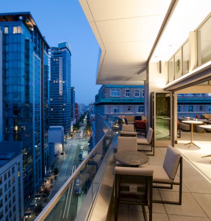 A modern urban balcony with seating overlooks a cityscape of glass buildings and a street below during the evening.