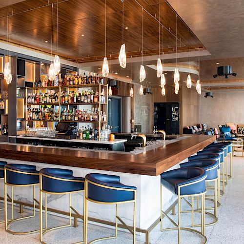 A modern bar with blue stools, pendant lights, and a well-stocked liquor shelf. There's seating in the background with a cozy atmosphere.