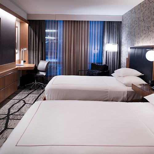A modern hotel room with two beds, a TV, desk, chair, and stylish decor. Curtains cover the window, and lighting is soft and ambient.