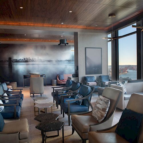 A modern lounge area with large windows, overlooking a cityscape and water, features comfortable seating and a mural on the back wall.
