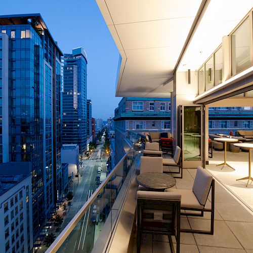 A modern balcony at dusk, with glass tables and chairs, overlooks a city street lined with tall buildings.