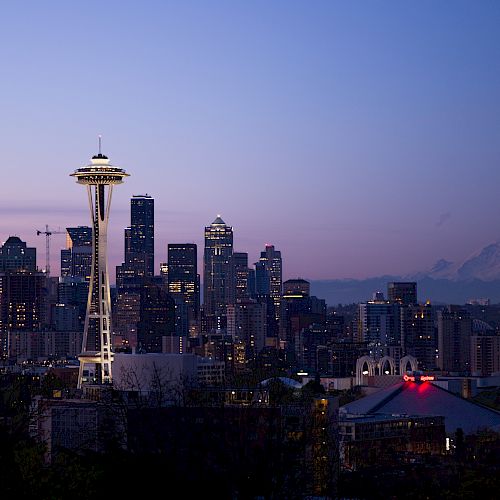 The image shows the Seattle skyline at dusk, featuring the Space Needle with Mount Rainier in the background under a twilight sky.