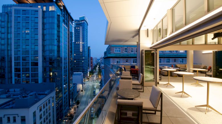 A modern balcony overlooking a cityscape at dusk, with tables and chairs, adjacent to tall glass buildings.
