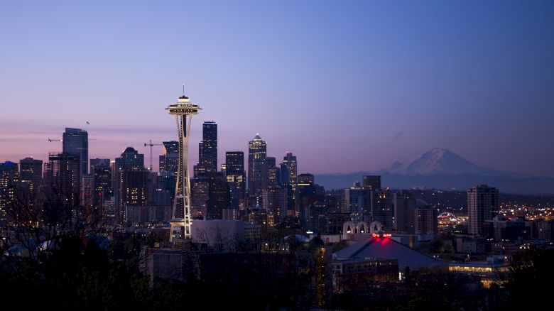The image shows the Seattle skyline at dusk, featuring the Space Needle, with mountains visible in the background.