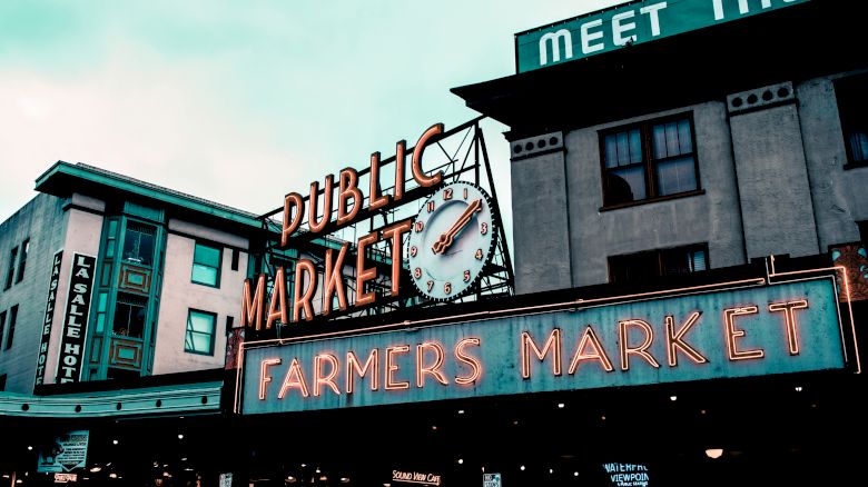 The image shows a neon sign for "Public Market" and "Farmers Market" with a clock, possibly Pike Place Market in a city setting.