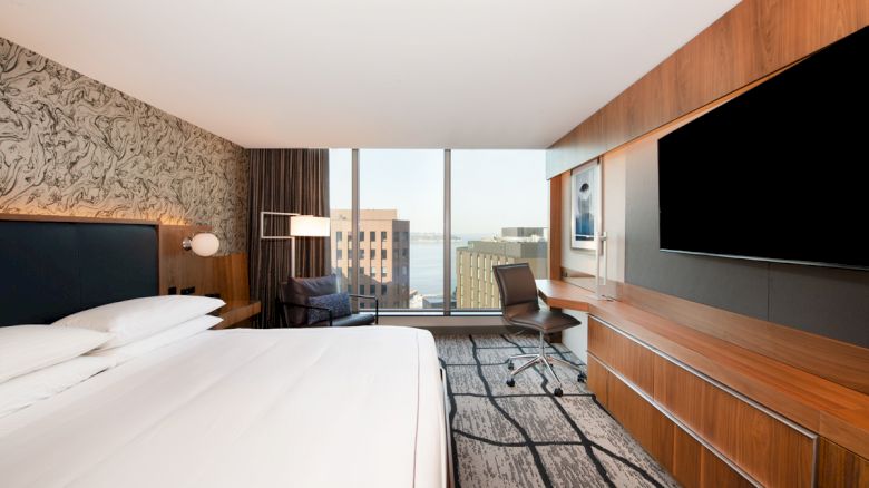 A modern hotel room with a large bed, work desk, and city view through the window, featuring contemporary decor and a wall-mounted TV.