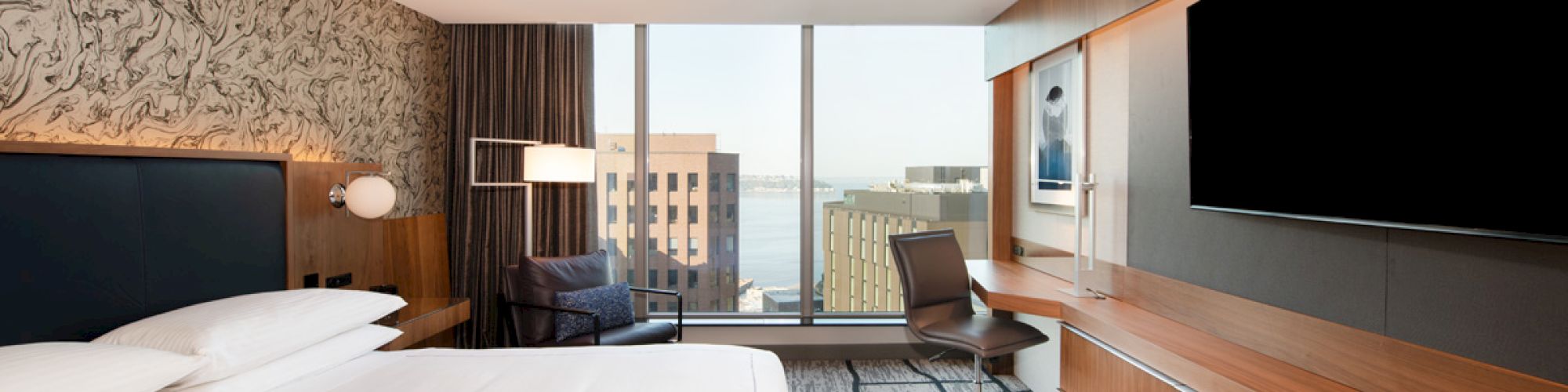 A modern hotel room with a large bed, desk, chair, flat-screen TV, and a window with a city view, featuring a textured accent wall.